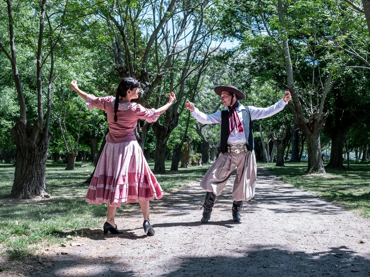 A couple in traditional Argentine gaucho and countryside dress dancing outdoors, arms raised (representative photograph)